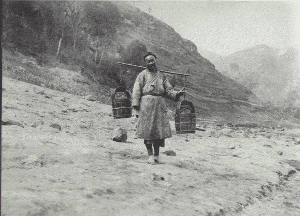 THE UBIQUITOUS WATER CARRIER

Drawing the water and hewing the wood are daily chores in China, mostly
carried out by women—though this is a picture of a man, a half-wit