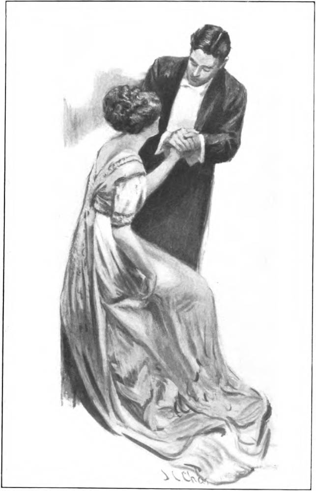 A smartly dressed man cups the hand of a seated woman in a long flowing dress.