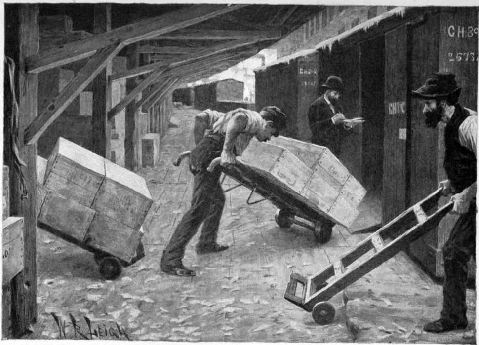 Three men are under cover on a loading dock.
There are box cars next to the loading dock.
One of the men is pushing a hand truck loaded with
crates into one of the boxcars.